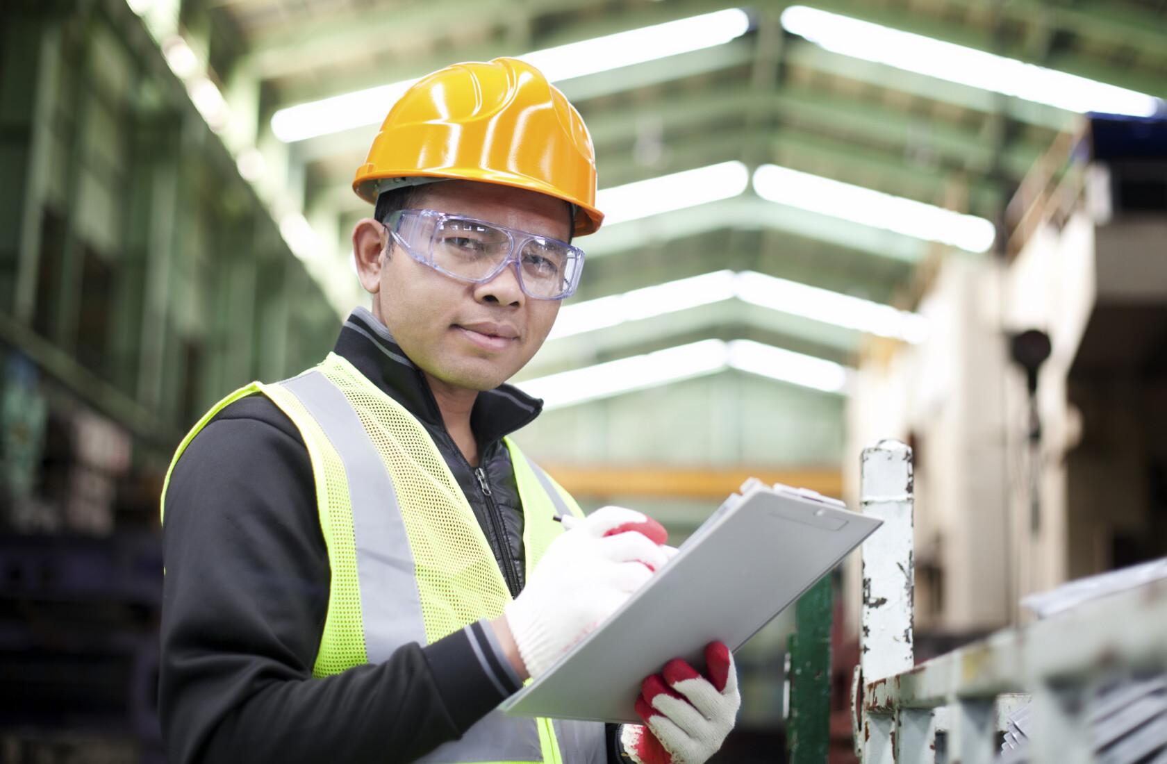 Man in a hard hat, safety vest, and protective glasses holds a clipboard and pen, standing in an industrial warehouse environment—ideal for consultations with lawyers in Chicago on workplace safety compliance.