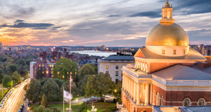 Boston State House with its golden dome illuminated at sunset, surrounded by trees and city buildings, stands near law offices offering litigation support, with cars and the river visible in the background.