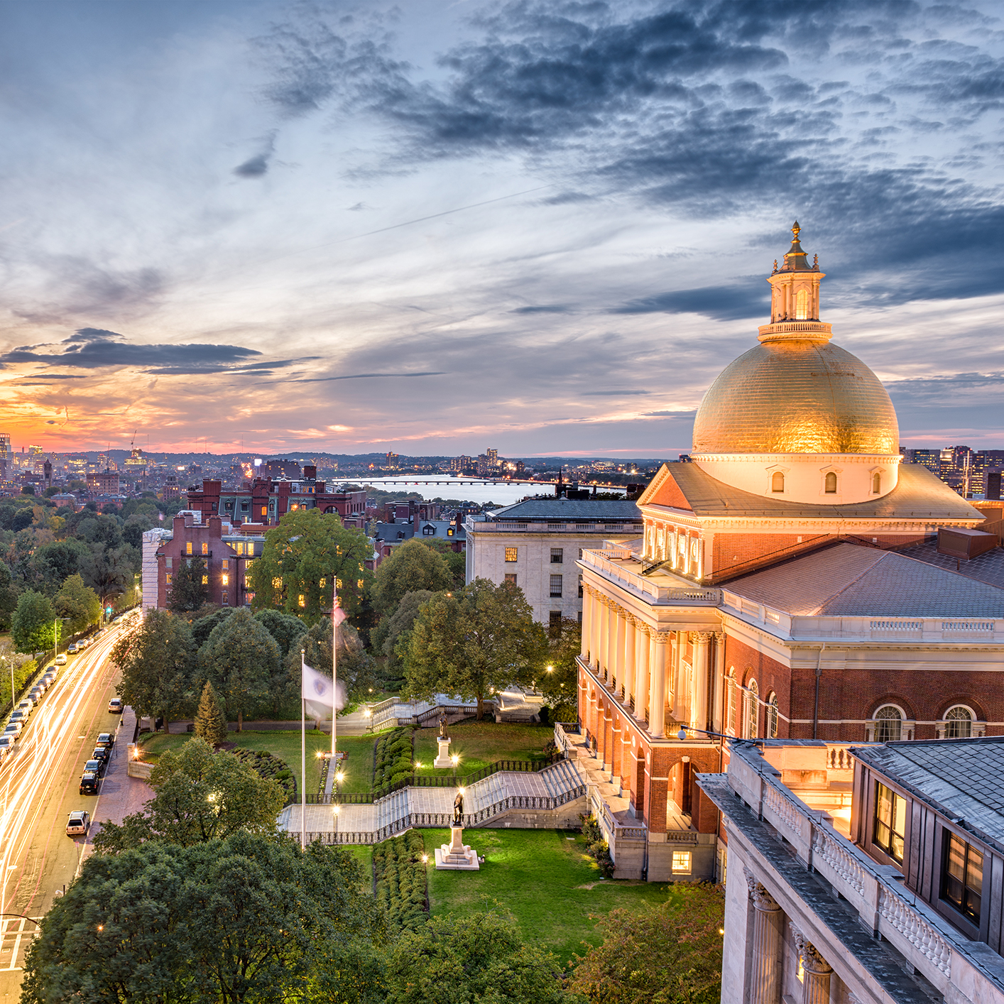Boston State House with its golden dome illuminated at sunset, surrounded by trees and city buildings, stands near law offices offering litigation support, with cars and the river visible in the background.
