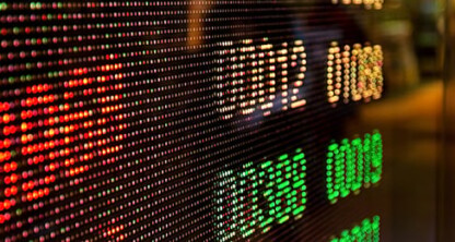 Una pantalla digital de la bolsa muestra filas de números y gráficos de colores, con una escena urbana difuminada al fondo, lo que refleja el trepidante ritmo del mundo exterior a un bufete de abogados corporativos.