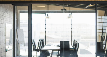 A modern glass-walled conference room with a long table, office chairs, and a flip chart, viewed from outside in an industrial-style corporate law office.