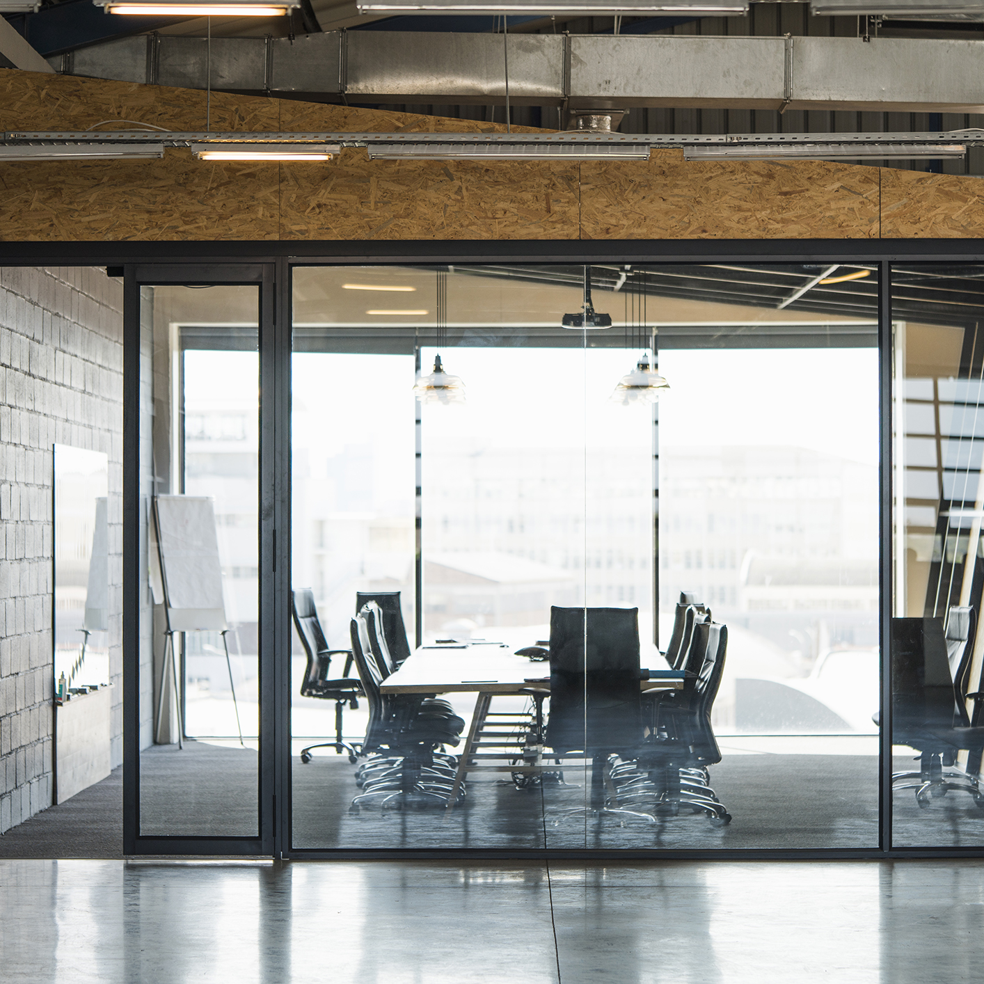 A modern glass-walled conference room with a long table, office chairs, and a flip chart, viewed from outside in an industrial-style corporate law office.