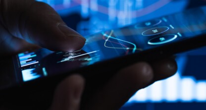 A hand holds a smartphone displaying financial graphs, with additional stock market charts visible on a screen in the background—ideal imagery for chicago lawyers seeking litigation support in complex financial or intellectual property law cases.