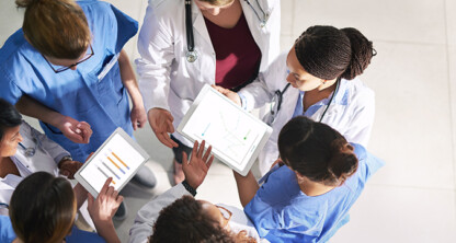 A group of healthcare professionals, wearing scrubs and lab coats, stand in a circle at a corporate law office and review data on digital tablets.
