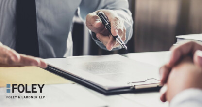 Dos personas están sentadas ante un escritorio en un bufete de abogados especializado en derecho mercantil; una de ellas sostiene un bolígrafo y señala un documento. El logotipo de Foley &amp; Lardner LLP aparece en la esquina inferior izquierda.