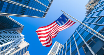 An American flag on a pole is centered among tall modern law offices, viewed from below against a clear blue sky, evoking the strength and integrity of a corporate law office.