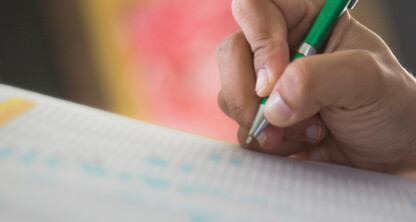 A person holds a green pen and writes on a sheet of paper with a chart or grid pattern, perhaps drafting documents in one of the law offices frequented by top chicago lawyers, with a blurred background.