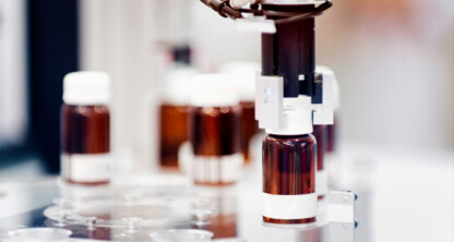 A robotic arm handles a brown glass vial with a white cap in a laboratory setting at one of the top law offices; several similar vials are visible in the background.