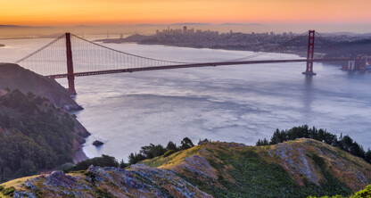 The Golden Gate Bridge spans across the water at sunset, with the city skyline and nearby law offices visible in the background and green hills in the foreground.