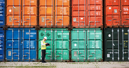 A worker in a safety vest and helmet stands in front of stacked colorful shipping containers, holding a clipboard and inspecting the area—much like lawyers in Chicago carefully review every detail for their clients.