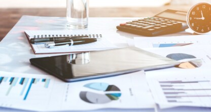 A desk in a corporate law office with financial charts, a notepad, a calculator, a glass of water, an analog clock, and a tablet, sunlight streaming through the window—an organized space ideal for litigation support.