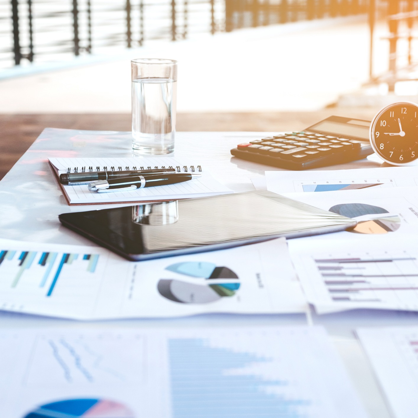 A desk in a corporate law office with financial charts, a notepad, a calculator, a glass of water, an analog clock, and a tablet, sunlight streaming through the window—an organized space ideal for litigation support.