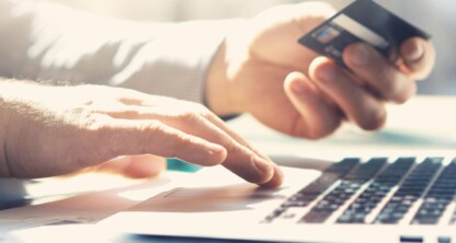 A person holds a credit card in one hand and types on a laptop keyboard with the other, possibly searching for law offices or chicago lawyers, with a smartphone lying nearby on the desk.