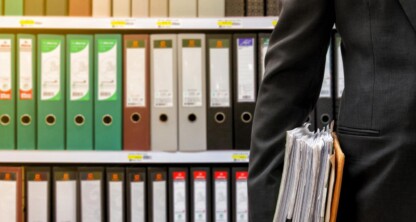 一名身穿西装的人手持一叠文件，身后的书架上整齐地摆放着各类活页夹和文件夹，生动展现了公司律师事务所中典型的严谨文件管理。