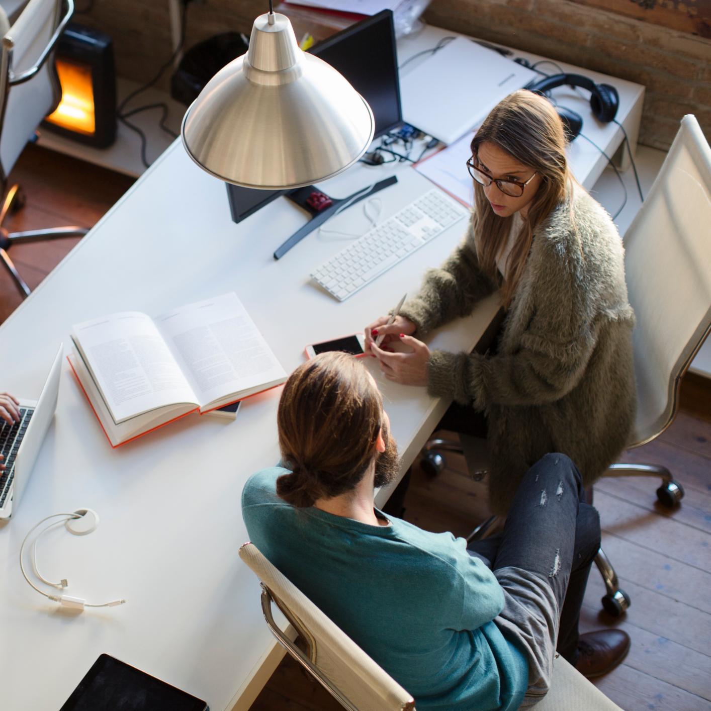 Two people sit at a white desk with laptops, an open book, and gadgets, engaged in conversation in a modern law office setting, reflecting the collaborative spirit of Chicago lawyers.