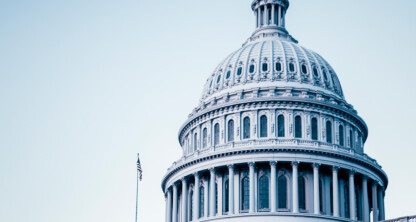 The image shows the dome of the United States Capitol building in Washington, D.C., with the American flag visible on the left side and a pale blue sky in the background—an iconic view often associated with law offices and legal institutions.
