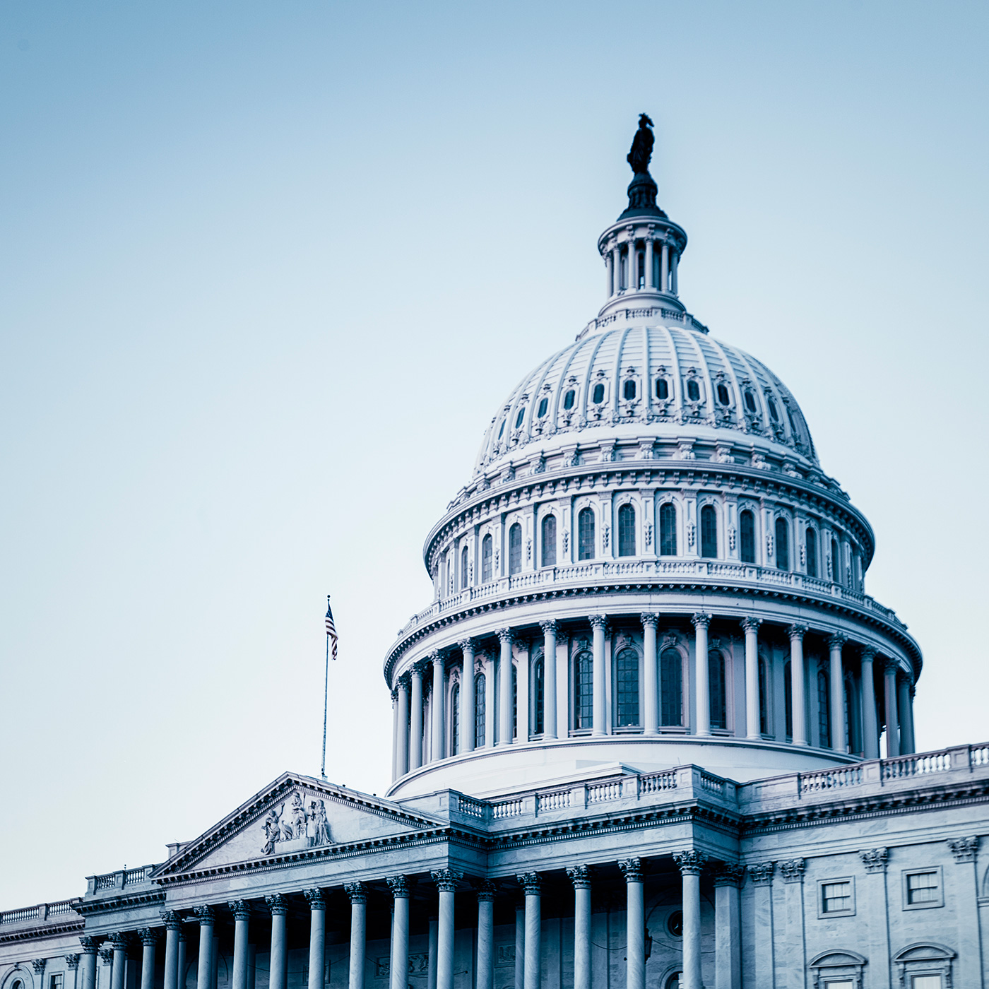 The image shows the dome of the United States Capitol building in Washington, D.C., with the American flag visible on the left side and a pale blue sky in the background—an iconic view often associated with law offices and legal institutions.