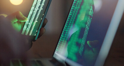 Person holding a smartphone displaying code next to a laptop screen with green code in a dimly lit setting, ideal for showcasing litigation support tools in a corporate law office.