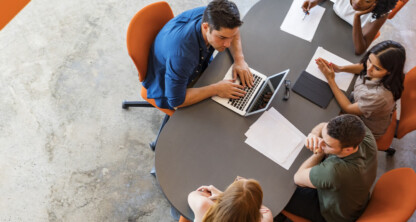 Four people sit around an oval table with papers, a laptop, and notepads, engaged in a meeting or group discussion about intellectual property law in a modern law office setting.