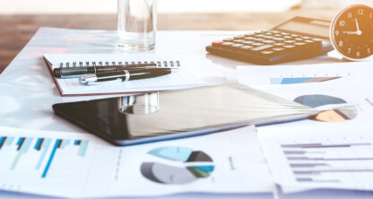 A desk in natural light with financial charts, a tablet, notepad, glass of water, calculator, and analog clock—an ideal setup often found in law offices or used by lawyers in Chicago handling intellectual property law cases.