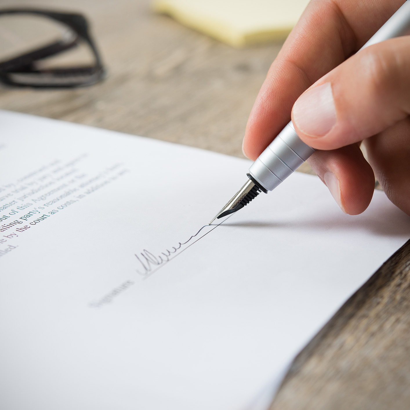 A hand holding a fountain pen signs a document on a wooden surface, with a pair of eyeglasses and sticky notes in the background—an everyday scene at a busy corporate law office in Chicago.