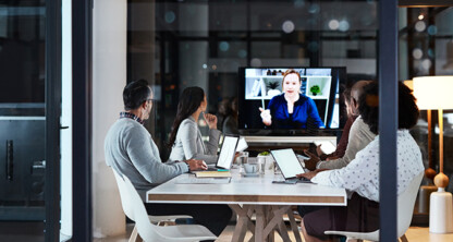 Four people sit at a conference table with laptops, as chicago lawyers join a video call with a woman displayed on a large screen at the front of the room.
