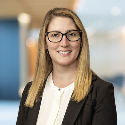 A woman with straight blonde hair and glasses, wearing a black blazer and white blouse, smiles in a corporate law office setting with a blurred background.