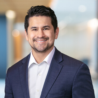 Man in a dark suit jacket and white shirt smiling, standing in a modern, blurred corporate law office—reflecting the professionalism of leading Chicago lawyers.