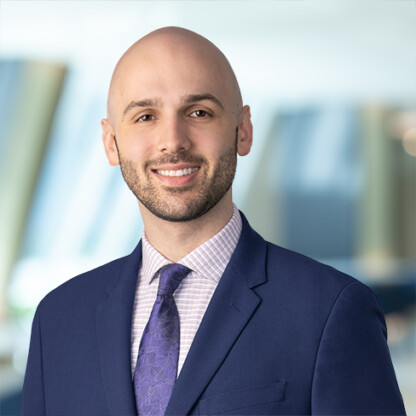 A smiling man with a bald head and trimmed beard, wearing a navy suit, patterned tie, and checkered shirt, stands confidently in a corporate law office setting.