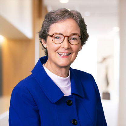A woman with short gray hair and glasses, wearing a blue blazer and white turtleneck, smiles while standing in a brightly lit corporate law office.