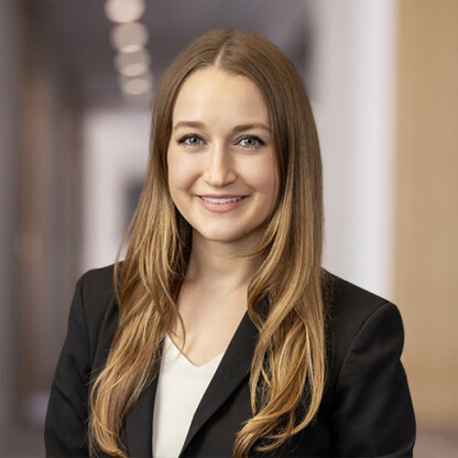 A young woman with long, light brown hair, wearing a black blazer and white top, smiles at the camera in a professional corporate law office setting.