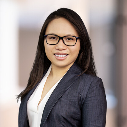 A woman with long dark hair and glasses, dressed in a dark blazer and white blouse, stands against a blurred indoor backdrop—reflecting the professionalism of Chicago lawyers in law offices.