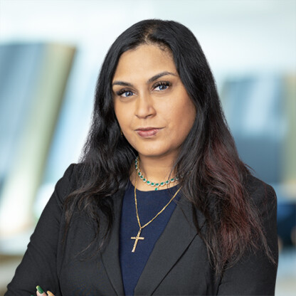 Woman with long dark hair wearing a black blazer and layered necklaces, including a cross pendant, stands in a blurred law offices setting in Chicago with arms crossed, embodying confidence and professionalism found among top lawyers in Chicago.