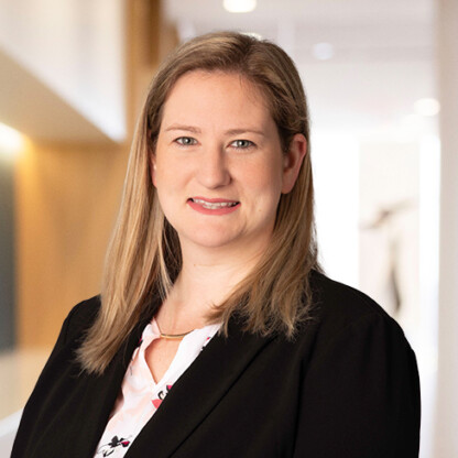 A woman with straight blonde hair wearing a black blazer and patterned blouse stands indoors in a corporate law office, facing the camera and smiling slightly.