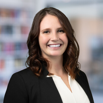 A woman with wavy brown hair wearing a black blazer and white blouse smiles at the camera in a blurred corporate law office, capturing the professionalism of Chicago lawyers.