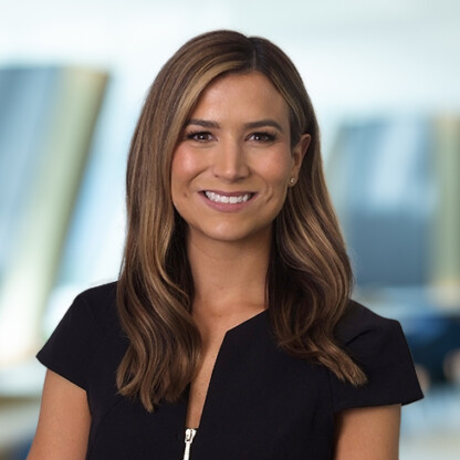 A woman with long brown hair, wearing a short-sleeve black top, smiles at the camera in a blurred corporate law office setting.