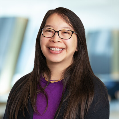Woman with long dark hair and glasses, wearing a purple top and black jacket, smiling in a bright, blurred corporate law office setting.