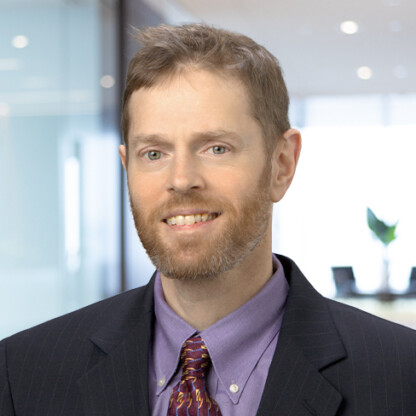 A man with light brown hair and a beard, wearing a dark suit, purple shirt, and patterned tie, is smiling in a corporate law office in Chicago, the blurred background hinting at a team of lawyers in Chicago.