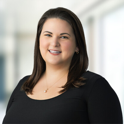 Woman with straight dark hair, wearing a black top and gold necklace, smiling in a well-lit corporate law office with a blurred background.