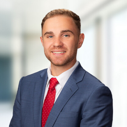 Man in a blue suit and red tie smiling, posed in front of a blurred corporate law office background.