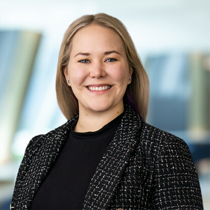 A woman with long blonde hair, wearing a black top and a black textured blazer, smiles at the camera against a blurred corporate law office background.