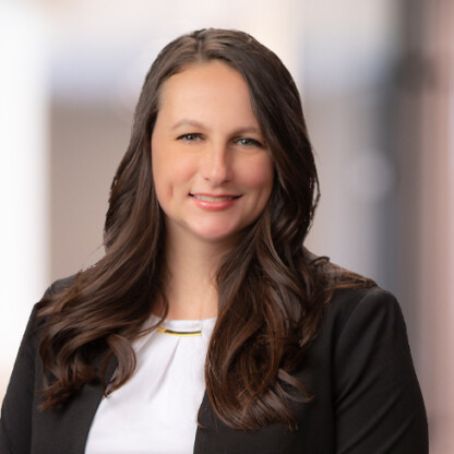 A woman with long brown hair wearing a black blazer over a white top, smiling at the camera against a blurred indoor background, often seen in law offices specializing in intellectual property law.