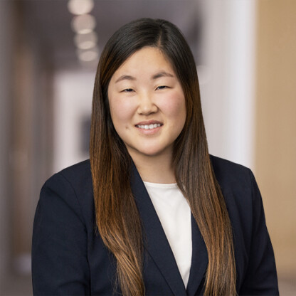 A woman with long straight hair wearing a navy blazer and white top, smiling, poses in a hallway with blurred lights in the background, reflecting the professional atmosphere found in law offices specializing in litigation support.
