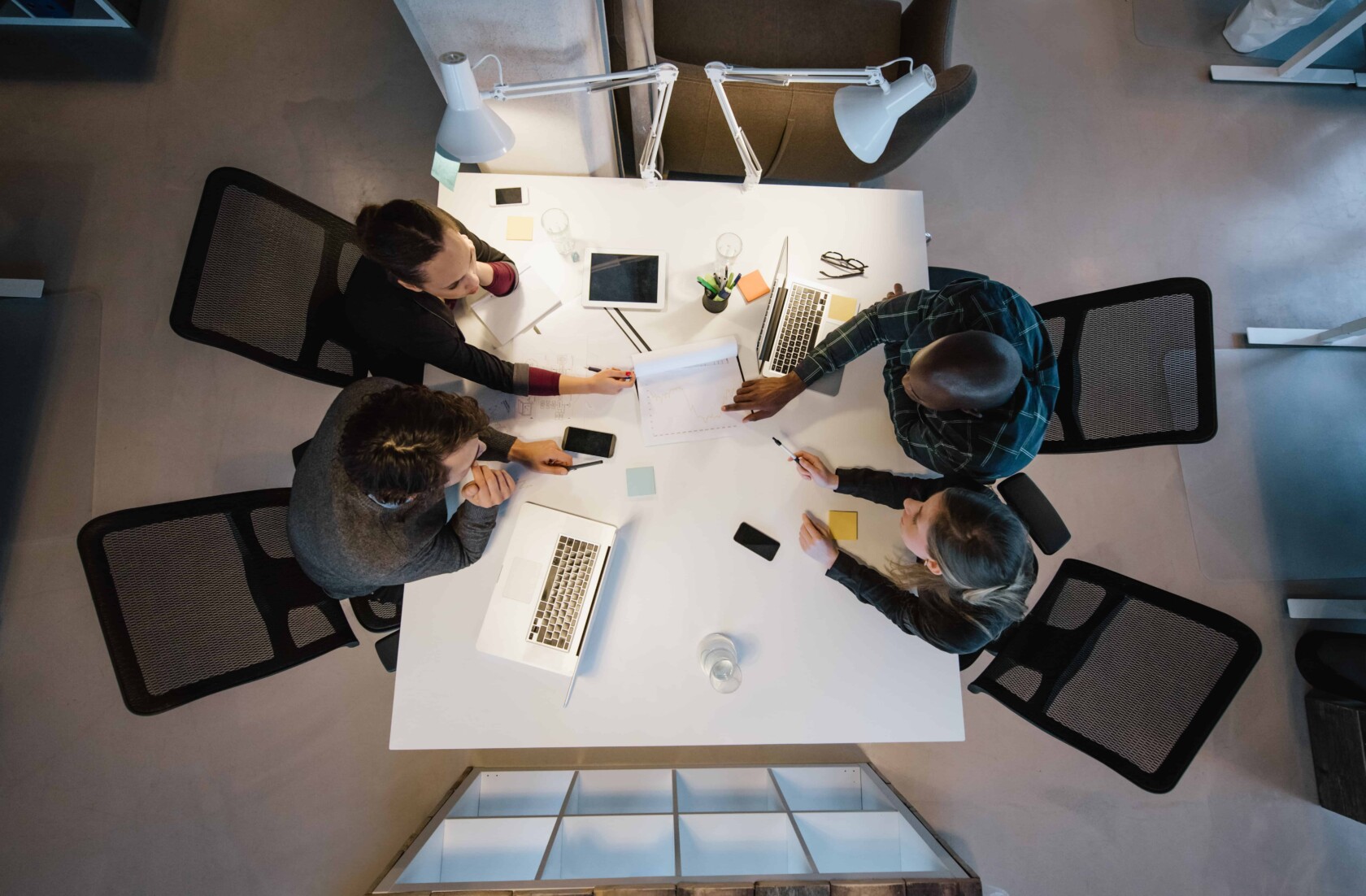 Four people sit around a white table with laptops, tablets, and papers, collaborating on work in a corporate law office setting.