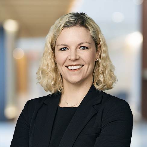 A woman with wavy blonde hair wearing a black blazer smiles at the camera, standing in a brightly lit corporate law office hallway.