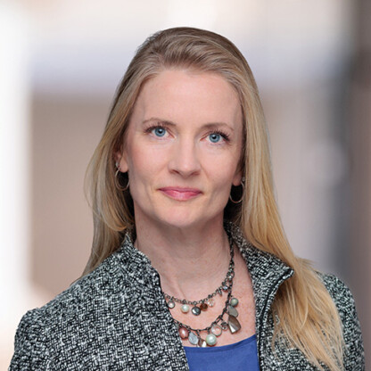 A woman with long blonde hair, wearing a gray textured jacket, blue top, and layered necklace, poses in front of a blurred indoor background at one of the leading law offices for Chicago lawyers.