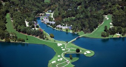 Aerial view of a golf course with fairways and greens extending onto a small peninsula, surrounded by water and wooded land, with nearby residential buildings and prominent law offices specializing in intellectual property law.