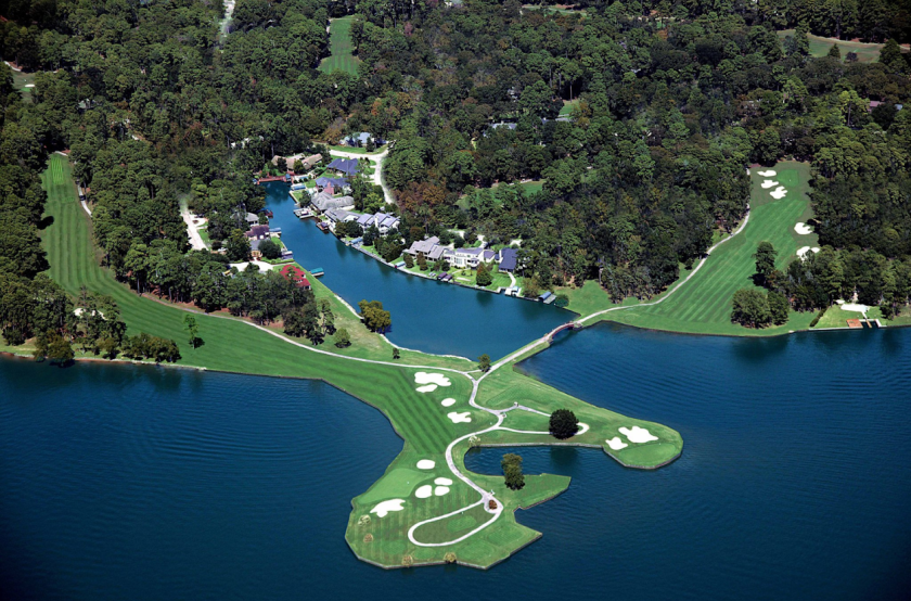 Aerial view of a golf course with fairways and greens extending onto a small peninsula, surrounded by water and wooded land, with nearby residential buildings and prominent law offices specializing in intellectual property law.