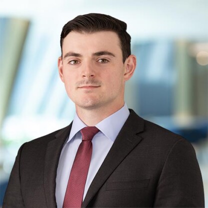 A man in a dark suit, light blue shirt, and red tie stands in front of a blurred corporate law office background, looking at the camera with a neutral expression.