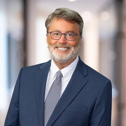 A middle-aged man with gray hair and beard, wearing glasses, a blue suit, white shirt, and striped tie, stands smiling in a blurred office hallway—reflecting the professionalism found in top law offices.